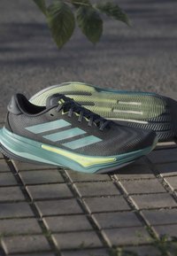 Gray athletic sneakers with light green accents and three striped design on the side. Textured upper and distinct sole pattern visible.