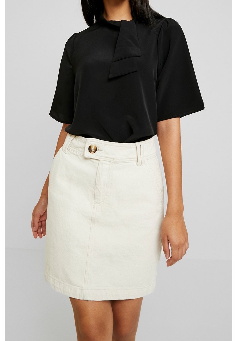 Femme portant un chemisier noir à manches courtes avec une cravate au cou et une jupe boutonnée taille haute couleur blanc cassé, debout devant un fond uni.