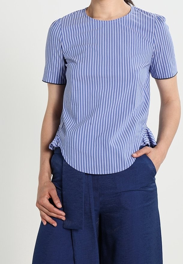 Femme portant un chemisier bleu et blanc à rayures verticales et manches courtes avec un pantalon large bleu marine, une main dans la poche.