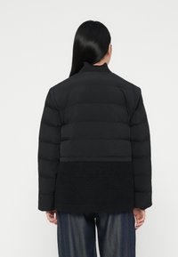 Melna pūkainā jaka ar horizontālu quilted dizainu, augstu apkakli un faktūru melnā apakšējā panelī. Modelis valkā brīvlaižu džinsu bikses.