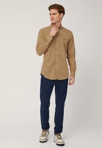 Camicia a maniche lunghe a quadri in senape con piccoli motivi geometrici, abbinata a jeans blu scuro e sneakers crema con dettagli neri.
