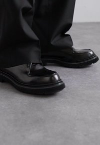 Black leather shoes with a glossy finish, square toe, and thick black rubber sole featuring ridges. Worn with black pants on a gray floor.
