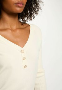 Top a coste color crema con scollo a V, caratterizzato da tre bottoni dorati lungo la scollatura e maniche lunghe.
