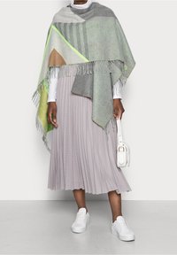 Personne portant un châle à franges gris-vert, une jupe plissée lilas, un haut à manches longues blanc, des baskets blanches, et tenant un petit sac à main blanc.