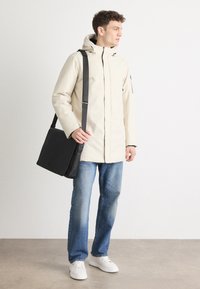 Beige vandtæt jakke med hætte, sort crossover taske, blå jeans og hvide sneakers. Glat tekstur og minimale design detaljer.