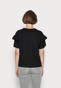 Schwarzes T-Shirt mit kurzen Ärmeln und Rüschen-Details, getragen mit grauen Jeans. Der Stoff wirkt weich und leicht.