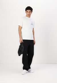 Balta trumpa rankovėmis marškinėliai su mėlynu piešinuku kairiajame krūtinės šone, derinami su juodais oversized kelnėmis ir baltais sportiniais bateliais. Laiko juodą maišą.