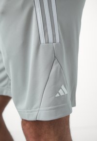 Gros plan sur un short de sport gris clair avec trois bandes verticales et un petit logo Adidas sur le côté inférieur près de la jambe d'un homme.