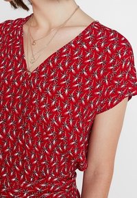 Červené šaty s krátkými rukávy, s výstřihem do V a bílým vzorem hvězdného výbuchu. Tkanina vypadá lehce a texturovaně. Viditelné zlaté vrstvené náhrdelníky.