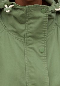 Zielona kurtka z kapturem o gładkiej, matowej fakturze, zapinana na napy, z dodatkowymi troczkami przy kołnierzu, umożliwiającymi regulację dopasowania.