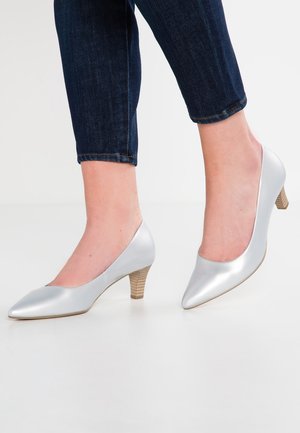 Person iført sølvfarvede kitten heel-sko med spidse tæer, stablede træhæle og mørke cropped denimjeans.