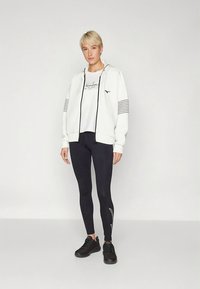 Sudadera blanca con cremallera y rayas negras, combinada con una camiseta gráfica blanca y mallas negras, completado con zapatillas deportivas negras.