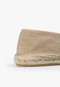 Vævet beige espadrille med et fiskebensmønster, rund tå og jute-sejladsdetaljer langs sålen. Flad, naturlig gummibund.