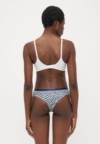 Woman seen from the back wearing a white bra with adjustable straps and zebra-print underwear with a dark waistband against a plain background.