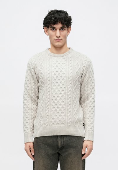 Pull en tricot gris clair avec un motif en câble texturé, des poignets et un ourlet côtelés, et des boutons sur les côtés. Porté avec un jean gris foncé.