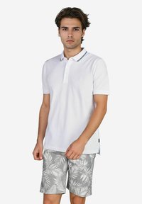 Weißes Polo-Shirt mit Kragen und zwei Knöpfen, mit einem marineblauen gestreiften Saum. Kombiniert mit grauen Shorts, die ein weißes Blättermuster aufweisen.