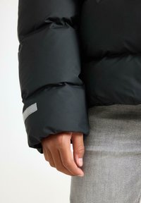 Veste matelassée noire avec une texture lisse et mate. Présente une manche ajustée et un accent réfléchissant près du poignet. Tissu gris visible en dessous.
