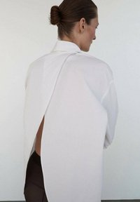 Camisa blanca de gran tamaño con un diseño de espalda abierta, que presenta hombros caídos y una tela suave y lisa. Lleva puesta sobre leggins oscuros.