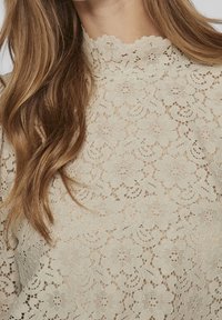 Primo piano di un top in pizzo floreale beige con scollatura a scallop e maniche lunghe, che mostra intricati delicati motivi floreali e texture.