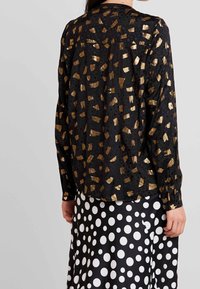 Blusa nera con motivi astratti dorati, maniche lunghe e vestibilità ampia. Indossata sopra una gonna a pois bianca e nera. Tessuto strutturato.
