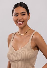 Beige gerippter Tanktop mit breiten Trägern und V-Ausschnitt, kombiniert mit einer goldenen, runden Diskus-Halskette. Glatte Textur und figurschmeichelndes Design.