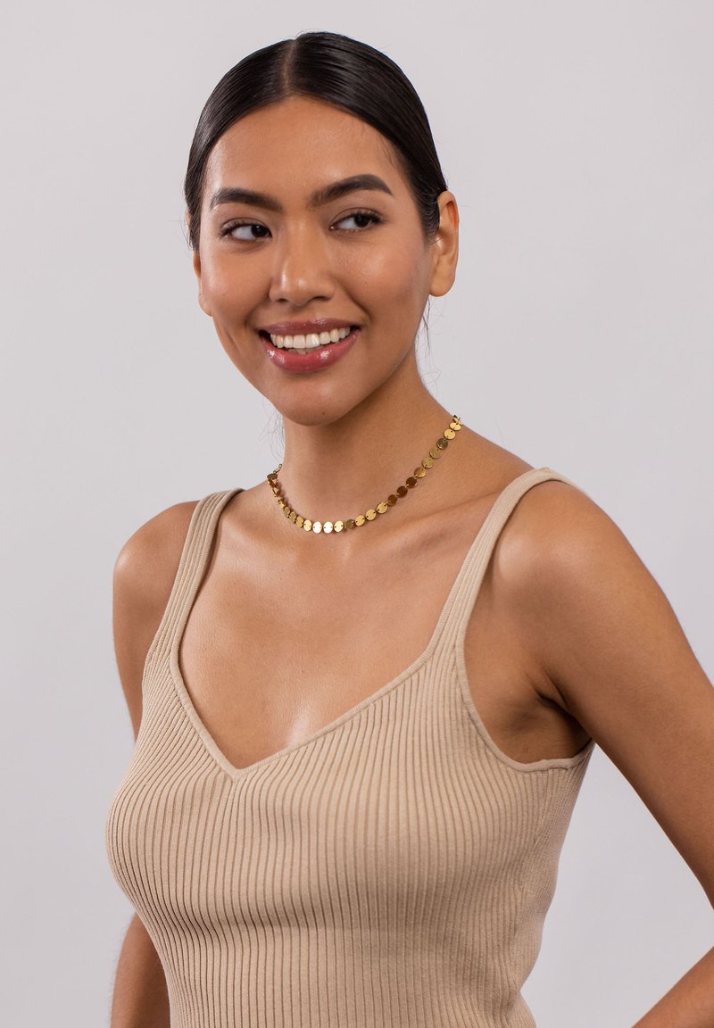 Beige gerippter Tanktop mit breiten Trägern und V-Ausschnitt, kombiniert mit einer goldenen, runden Diskus-Halskette. Glatte Textur und figurschmeichelndes Design.