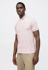 Polo rosa claro com gola, mangas curtas e acabamento branco. Apresenta um pequeno emblema com logotipo no peito. Textura suave.