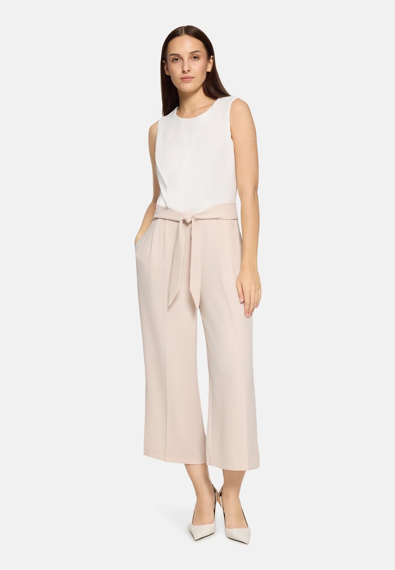 Haut blanc sans manches associé à un pantalon large beige clair, avec une taille nouée. Le modèle porte des talons pointus de couleur neutre.