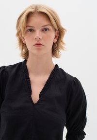 Schwarze Leinenbluse mit einem rüschierten V-Ausschnitt und kurzen, puffigen Ärmeln mit gerafften Details. Einfache, glatte Textur und keine Muster.