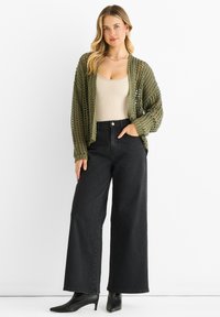 Olive green open-knit cardigan over a beige fitted top, paired with high-waisted black wide-leg jeans and black pointed ankle boots.