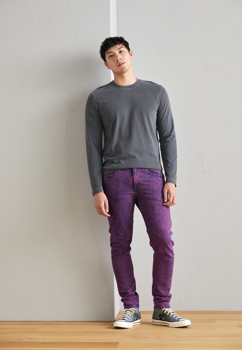 Camisa de manga larga gris combinada con jeans morados con un patrón texturizado; modelo con zapatillas grises y blancas de pie contra una pared.