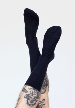 Schwarze gerippte Socken, die auf einem Hintergrund präsentiert werden. Der Stoff wirkt weich, hat eine glatte Textur und schmiegt sich eng um die Knöchel.
