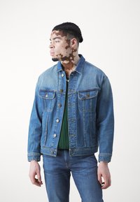Denim jakna u svijetloplavom do srednje plavom gradientu s dva prednja džepa, metalnim gumbima i oštrim ovratnikom, složena preko zelene rebraste puloverice.