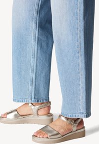 Sandales argentées métalliques avec une large sangle, une boucle à la cheville et une semelle épaisse beige, associées à un pantalon en denim bleu clair à jambes larges.