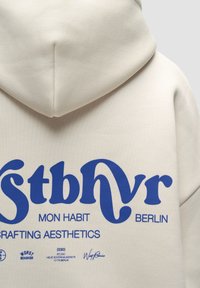 Cremfarbener Kapuzenpullover mit blauem Grafikdesign und dem Text: "stbvr MON HABIT BERLIN CRAFTING AESTHETICS" sowie markenspezifischen Akzenten.