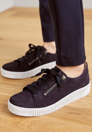 Zwarte suède sneakers met dikke witte rubberen zolen, zwarte veters en zijritsen, gedragen met korte zwarte broek op een houten vloer.