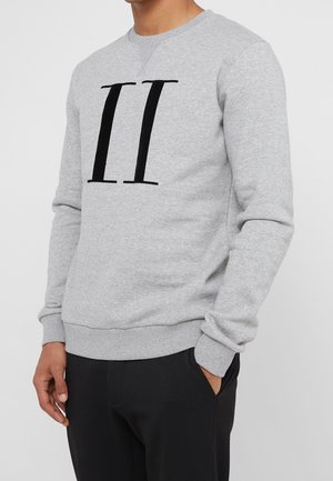 Uomo che indossa una felpa grigio chiaro con un grande numero romano nero II sul davanti e pantaloni neri, con una mano in tasca.