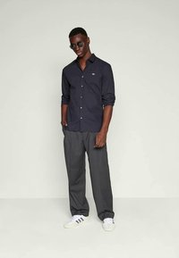 Homem em camisa escura de botões, calças largas com riscas, sapatilhas brancas e óculos de sol, de pé contra um fundo claro e simples.