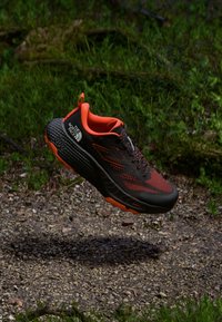 Czarne i pomarańczowe buty do biegania po szlakach z siateczkową cholewką, teksturowaną gumową podeszwą oraz charakterystycznym detalem logo. Posiadają wyściełany kołnierz i sznurowadła.