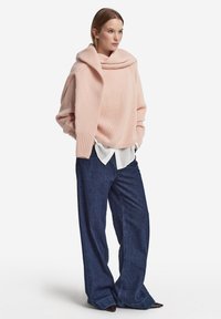 Rosa stickad tröja med en vid tubhals, lös passform, ribbade muddar; kombinerad med lösa mörka denimbyxor och en vit skjorta under.