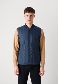 Colete acolchoado azul-marinho com costuras verticais, fecho de zip, gola e um bolso frontal, usado sobre uma camisa de mangas compridas bege.