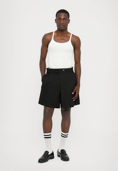 Schwarze Shorts mit gefälteltem Vorderteil, Seitentaschen und kontrastierenden Nähten. Kombiniert mit einem weißen Tanktop, schwarzen Slippern und gestreiften Socken.