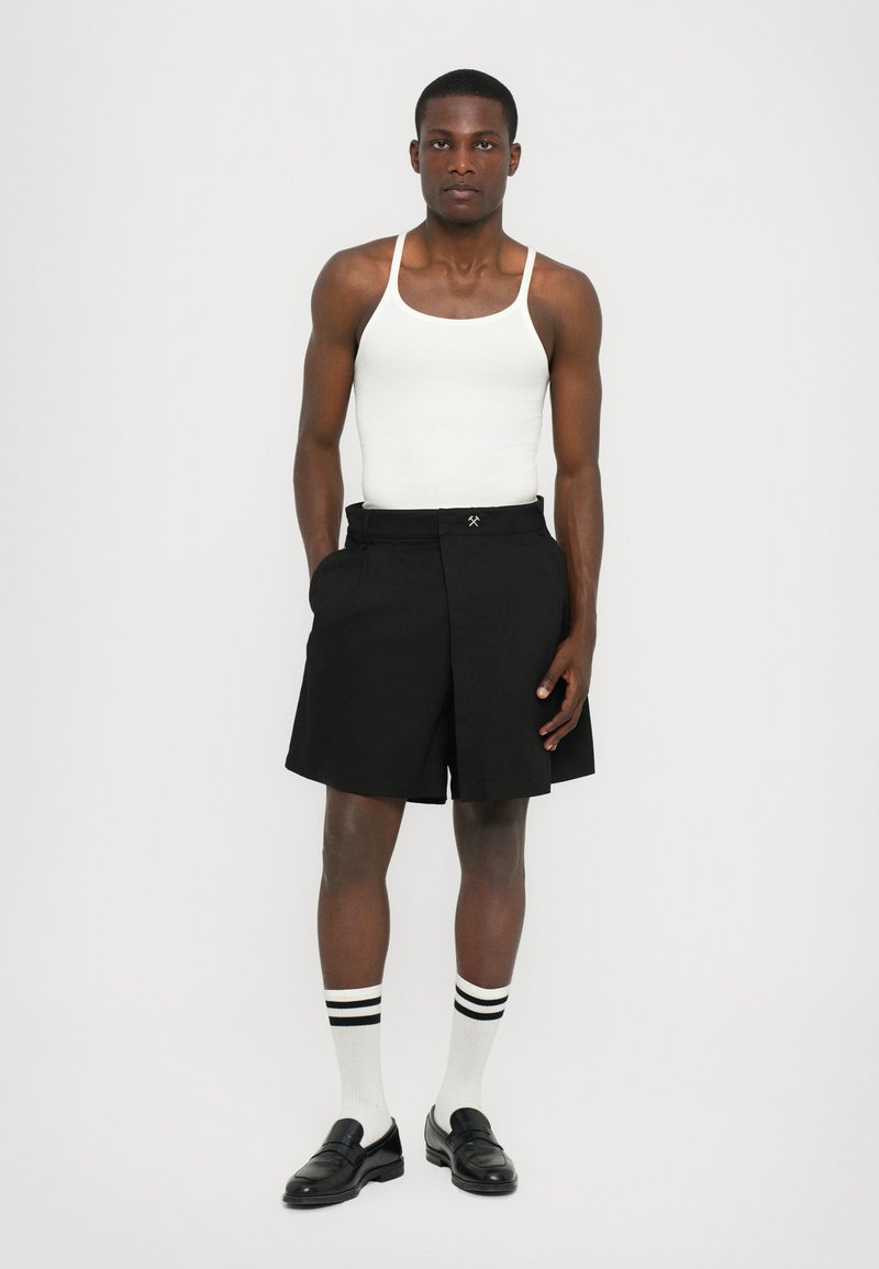Sorte shorts med plisseret front, sidelommer og kontrastsyning. Sammen med en hvid tanktop, sorte loafers og stribede sokker.