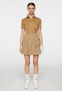 Brązowa koszulka polo z wzorzystym kołnierzykiem, w połączeniu z beżową spódnicą plisowaną. Białe sneakersy i biała rękawiczka golfowa dopełniają wygląd.