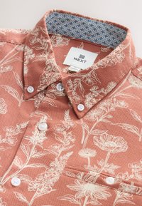 Chemise à motif floral corail avec imprimés botaniques blancs, présentant un tissu texturé, une fermeture à boutons, et un col intérieur bleu à motif.