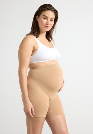 Έγκυος γυναίκα που φοράει λευκό αθλητικό σουτιέν και γυμνά σορτς υποστήριξης εγκυμοσύνης, στέκεται μπροστά σε απλό ανοιχτό φόντο.