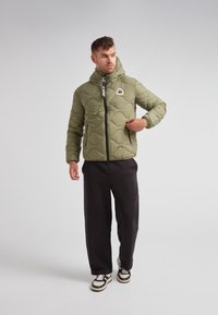 Padded olive green jacket with a hood, featuring a black zipper, elastic cuffs, and a triangular logo on the chest. Worn over black pants.