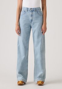 Γυναίκα που φοράει παντελόνι τζιν γαλάζιο, με ψηλή μέση και φαρδιά παντελόνα, συνδυασμένο με καφέ και κίτρινα αθλητικά παπούτσια και λευκό τοπ.