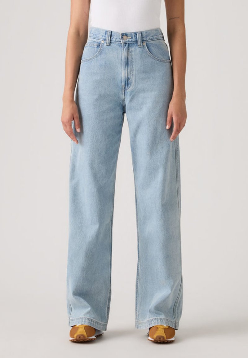 Γυναίκα που φοράει παντελόνι τζιν γαλάζιο, με ψηλή μέση και φαρδιά παντελόνα, συνδυασμένο με καφέ και κίτρινα αθλητικά παπούτσια και λευκό τοπ.