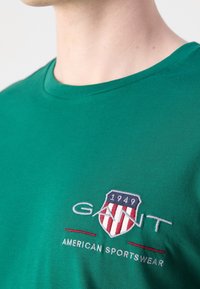 Zielona bawełniana koszulka z okrągłym dekoltem, z haftowanym herbem w kolorach czerwonym, białym i niebieskim oraz napisem "GANT AMERICAN SPORTSWEAR".
